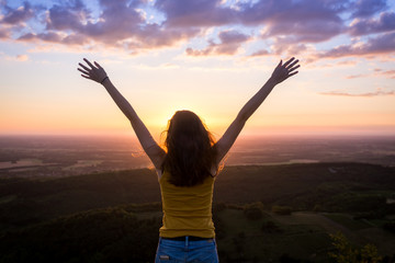 une jeune femme de dos , levant les bras face à un paysage sous le coucher de soleil