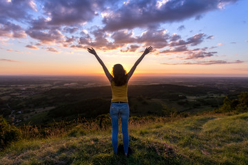 une jeune femme de dos , levant les bras face à un coucher de soleil, en haut d'une montagne