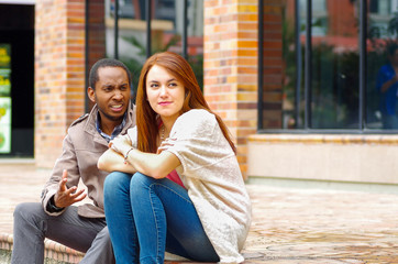 Fototapeta premium Interracial happy charming couple sitting on steps in front of building interacting and smiling for camera