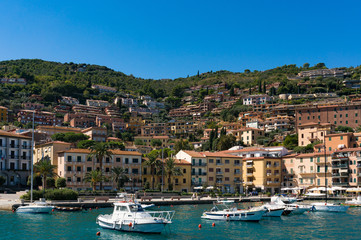 Fototapeta na wymiar Porto Santo Stefano buildings and bay with fishing boats and yachts on a sunny summer day. Mediterranean Italian resort village of Tyrrhenian Sea with luxury property on the background