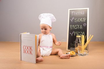 A little boy in a cap and apron cook cookbook studies. Beside him stand menu, spaghetti and colorful pasta