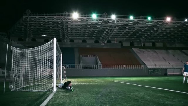 Soccer player scoring a goal into the net and goalkeeper falling down while trying to save the goal at the stadium in the night