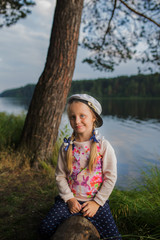 cheerful girl in a cap with two braids sits on a log by the river on a summer evening on the town