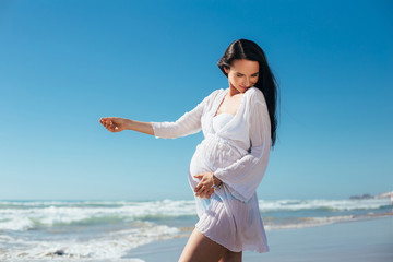 Fototapeta na wymiar Pregnant having fun on the beach. Expectant mother in white dress on a background of blue sky. Pregnant wearing a tunic seashore walk.