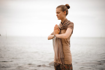 Fototapeta na wymiar Woman standing a grateful namaste yoga pose on the beach next to the ocean in cloudy weather. Zen, meditation, peace.