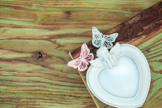 Heart shaped white photo frame with porcelain cupid angel and butterfly pins on wooden background