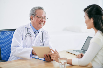 Obraz premium Vietnamese mature doctor with tablet computer talking to patient