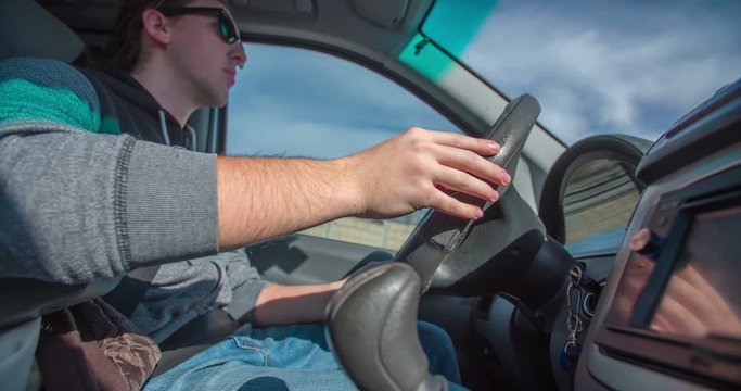 A young man is driving a van and he is tapping his figers to the music on a steering wheel. He is also preparing to change the gear. Close-up shot.
