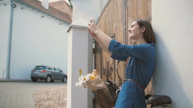 Brunette girl taking selfie, standing near old building with a bike with flowers in a basket, slow mo