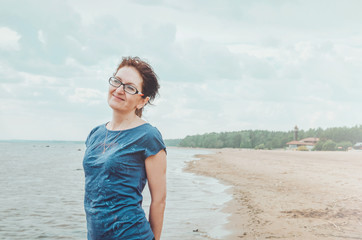Feminine portrait of attractive mature healthy woman enjoying happy on summer beach. Smiling female standing seaside with cloudy sky, blue baltic sea views, outdoors. Nature background, copy space.