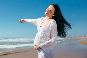 Fototapeta na wymiar Expectant mother in white dress on a background of blue sky. Pregnant wearing a tunic seashore walk. Pregnant having fun on the beach.