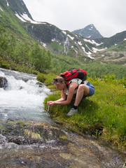 Young woman with backpack drinking from a stream on a background of mountains with snow
