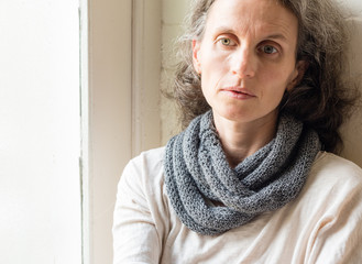 Middle aged woman in grey scarf next to window looking sad and thoughtful