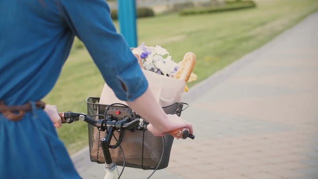 Girl hands on a handlebar of a bicycle, flowers and bread in a basket, cycling in the city, slow mo, steadicam shot