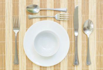 Closeup white ceramic dish and cup with stainless fork and spoon on wood mat textured background on dining table in top view , group of tableware before dinner