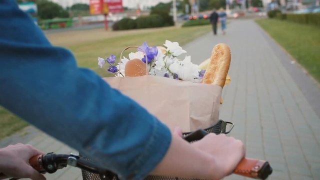 Woman hands on handlebar of bike with flowers and bread in basket as she walks in the street, slow mo, steadicam shot