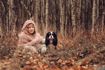 Naklejka na ściany i meble child girl relaxing with her cavalier king charles spaniel dog in autumn forest, wrapped in cozy scarf