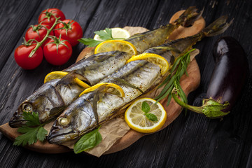 grilled mackerels on the black wooden table with lemon, tomatoes, eggplant, and herbs