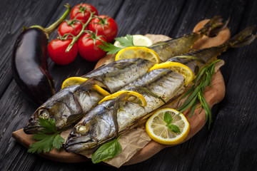 grilled mackerels on the black wooden table with lemon, tomatoes, eggplant, and herbs