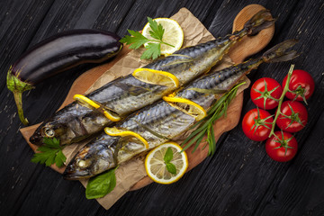 grilled mackerels on the black wooden table with lemon, tomatoes, eggplant, and herbs