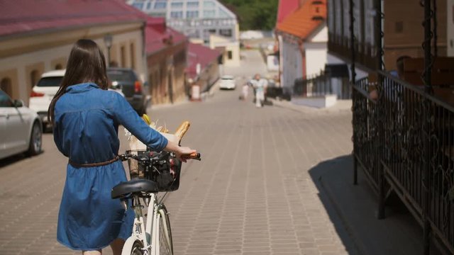 Back view of a girl with waving hair walking her bike with flowers and bread in a basket, 4k steadicam shot