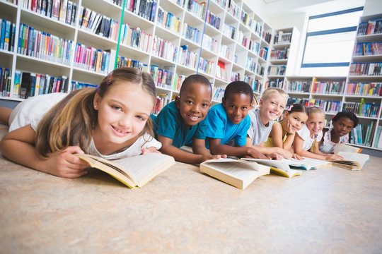 Smiling school kids lying on floor reading book in library 