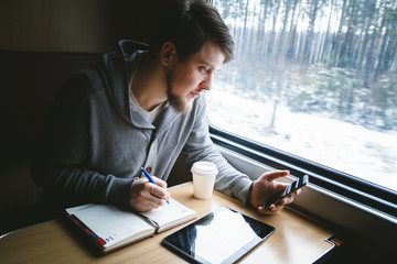 The man sits at a table in a train, he writes in a notebook and looking at the phone