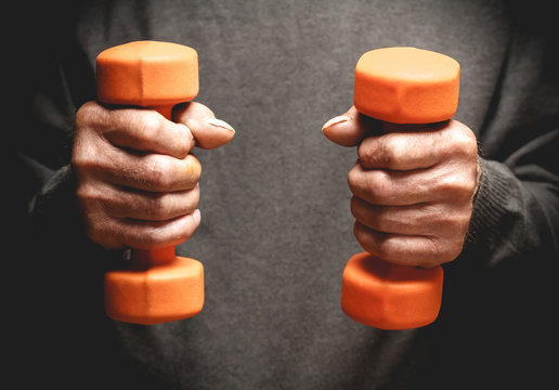 Orange dumbbells in the hands of an elderly man. Sports retired. Photo in low key