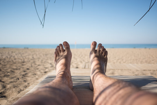 Men's feet on lounge first person view from bungalow on the sea with sand beach