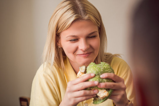 Picture of blond woman eating delicious vegan burger in vegan restaurant or cafe. Beautiful lady in yellow t-shirt havig lunch or dinner.
