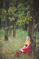 Fototapeta na wymiar Portrait of young beautiful blonde woman wearing a long red elegant dress posing in a green meadow. Fashionable sexy attractive girl with blue eyes in field. Gorgeous fair hair female, outdoor shot.