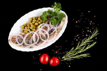 Slices herring with peas, decorated by onion on white plate on black background, top view