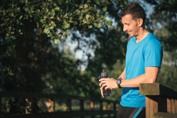 Runner man holding water bottle while resting during outdoor training workout. Fit fitness sport model leaning on wooden railing for motivation.