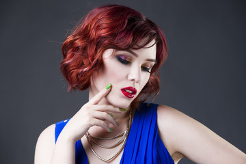 Young beautiful red-haired caucasian woman in blue dress posing in studio on gray background, professional makeup and hairstyle