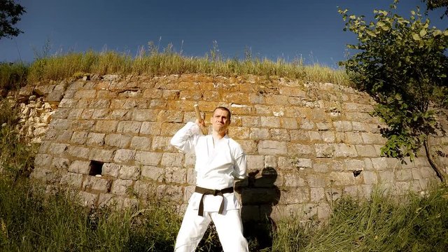 Man in white kimono trains with nunchaku with serious face background of wall