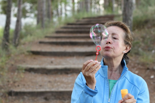 Senior woman blowing soap bubbles in summer park with copy space 