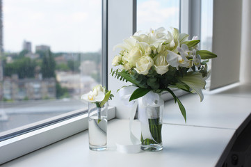 Beautiful wedding bouquet in a glass vase. Flowers on white windowsill. Boutonniere in a small vase. The window in the background.