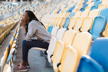 Side view of sad african american young woman sitting at stadium with her chin resting on hands and...