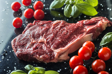 Fresh steak served with spices, tomatoes and leafs of basil on marble background. Uncooked beefsteak cooking on a kitchen. Delicious, spicy, juicy meat with copy space closeup.
