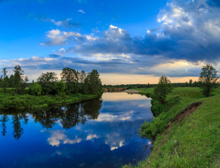 Sky with light clouds reflected in the river, field, trees, summer evening