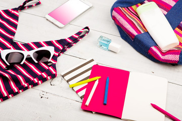 colorful striped bag, swimsuit, smart phone, sunglasses and notepads on white wooden desk