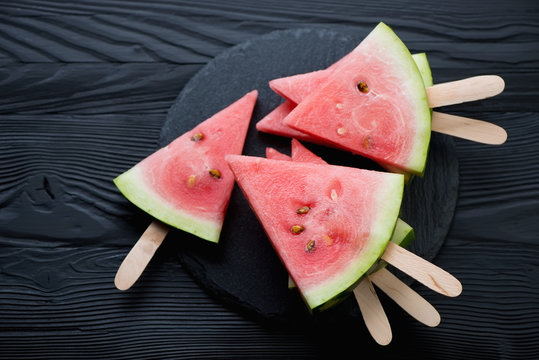 Watermelon slices on sticks, black wooden background, above view
