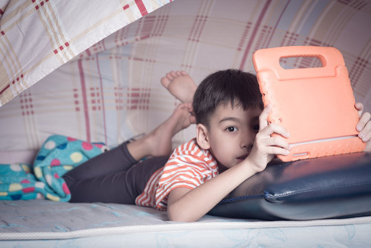 Little boy hinding under blacket playing tablet