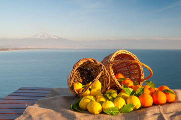 Wicker baskets full of oranges and lemons on a piece of jute with blue sea and Mount Etna in the background - 116346350