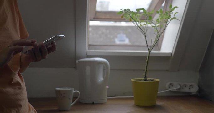 Woman using mobile phone at home while waiting for the kettle to boil the water for tea