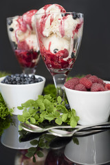 Fresh fruits raspberries and blueberries in white bowls and ice cream with the same fruits and raspberries syrup in glass. Stay on mirror table on black background