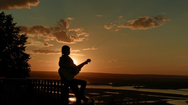 Young romantic woman playing the acoustic guitar, at sunset near camping on high hill, silhouette, wide angle