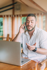 Young man sitting at cafe working on laptop and talking on mobile phone