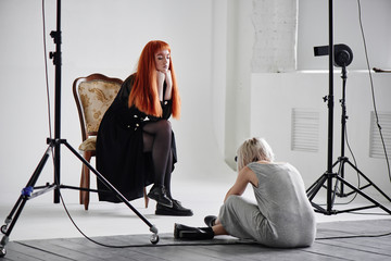 Girl photographer photographing fashion model in black sitting on a chair on white background in...