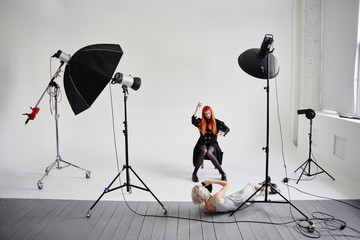 Girl photographer lies on the floor and shoots of fashion model posing on chair on white background...